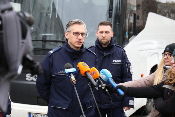 Policjanci podczas briefingu prasowego  na placu przy komendzie udzielają wywiadu. W tle stoi autokar