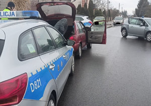 Radiowozy policji oraz bordowy ford stoją na drodze w tle zabudowania i policjaci