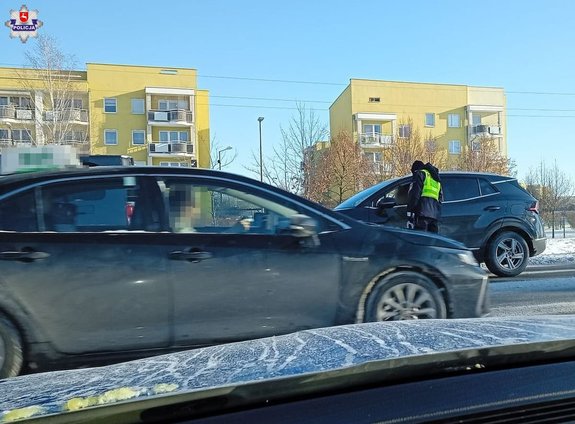 Policjant podczas kontroli trzeźwości na pierwszym i drugim planie dwa pojazdy osobowe a w oddali zabudowani miejski - bloki