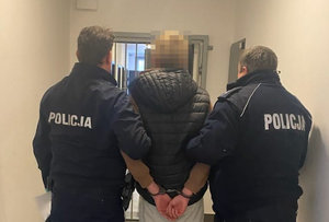 Zatrzymany mężczyzna z założonymi kajdankami jest prowadzony przez dwóch umundurowanych policjantów korytarzem
