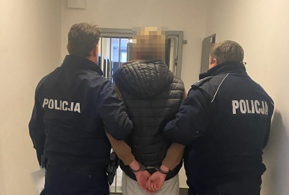 Zatrzymany mężczyzna z założonymi kajdankami jest prowadzony przez dwóch umundurowanych policjantów korytarzem