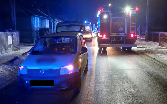 Zdjęcia z miejsca zdarzenia drogowego. widoczna jest ulica o nawierzchni asfaltowej na której stoją pojazdy policji i straży z włączonymi światłami błyskowymi oraz samochó osobowy marki fiat w tle zabudowa miejska. Zdjęcie w porze nocnej.