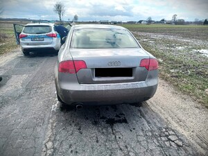 Pojazd marki Audi koloru szarego przedni stoi radiowóz oznakowany i policjant. Pojazdy stoją na asfaltowej drodze w tle zabudowania i pola uprawne,