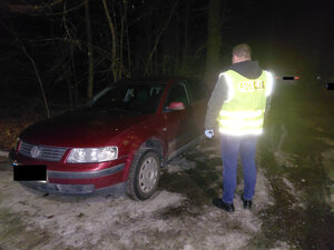 Policjant w kamizelce odblaskowej stojący obok bordowego volkswagena. Zdjęcie w porze nocnej. W tle drzewa i inne pojazdy zaparkowane.