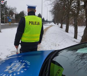 Na pierwszym planie widoczny jest przód radiowozu oznakowanego a obok niego stoi odwrócony plecami policjant umundurowany w kamizelce odblaskowej w tle widoczna jest droga i chodnik pokryty śniegiem