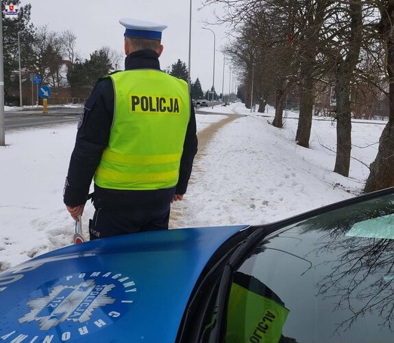 Na pierwszym planie widoczny jest przód radiowozu oznakowanego a obok niego stoi odwrócony plecami policjant umundurowany w kamizelce odblaskowej w tle widoczna jest droga i chodnik pokryty śniegiem