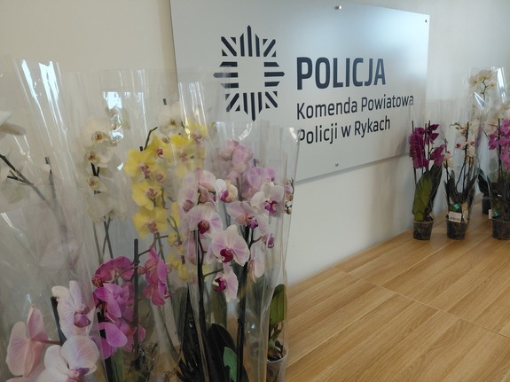 Kwiaty ustawione na stole w tle tablica z nazwą Komenda Powiatowa Policji w Rykach