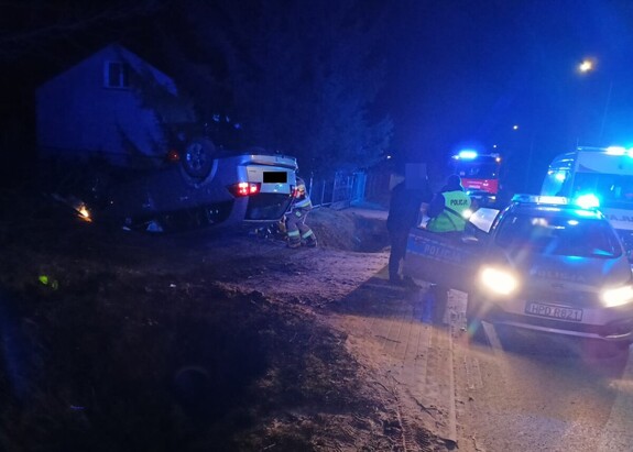 Samochód osobowy leżący na dachu uszkodzony w wyniku zdarzenia drogowego obok na drodze stoją pojazdy służb policji, straży i pogotowia ratunkowego. Policjant wraz z mężczyzną stoi obok radiowozu a strażacy przy srebrnym samochodzie.