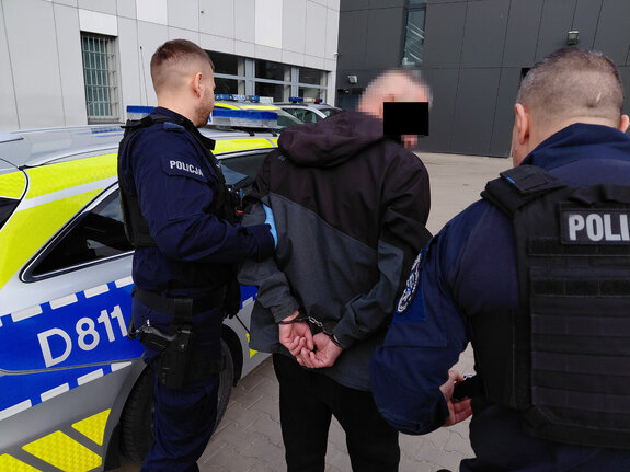dwóch umundurowanych policjantów prowadzi do radiowozu zatrzymanego mężczyznę z założonymi na ręce trzymane z tyłu kajdankami. W tle budynek komendy i radiowozy