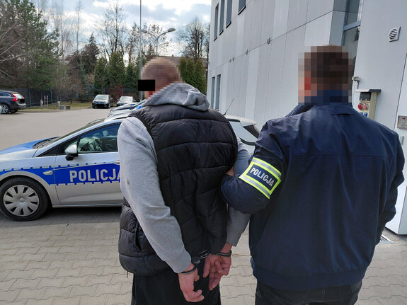 Zatrzymany mężczyzna z kajdankami założonymi na ręce z tyłu jest prowadzony przez policjanta w tle radiowozy i inne pojazdy zaparkowane na parkingu przy szarym budynku