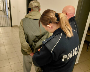 Policjantka zakłada zatrzymanemu mężczyźnie kajdanki na ręce trzymane z tyłu obok stoi jeszcze jedne policjant.