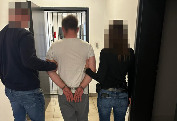 Policjant i policjantka nieumundurowani prowadzą korytarzem zatrzymanego mężczyznę z założonymi na ręce z tyłu kajdankami