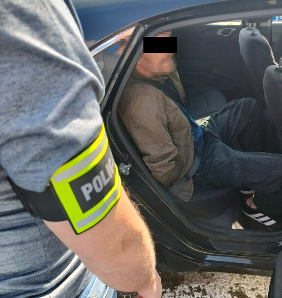 Zatrzymany mężczyzn siedzi w nieoznakowanym radiowozie obok pojazdu stoi policjant nieumundurowany z opaską z napisem policja