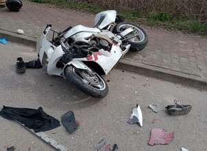 Motocykl koloru białego leżący na jezdni z widocznymi uszkodzeniami. Wokół pojazdu na asfaltowej nawierzchni leżą oderwane elementy pojazdu oraz but.