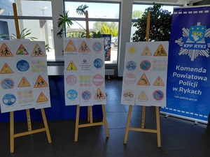 Prace dzieci przedstawiające znaki drogowe wystawione na trzech sztalugach w holu komendy obok baner reklamowy komendy policji w rykach