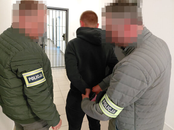 Policjant zakłada kajdanki na ręce trzymane z tyłu zatrzymanemu mężczyźnie drugi policjant stoi obok.