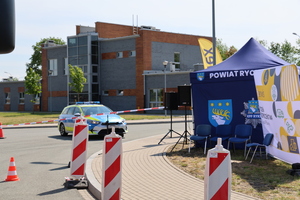 Radiowóz policyjny obok namiotu promocyjnego komendy policji w rykach, na asfalcie rozstawione są pachołki i taśma zabezpieczająca, w tle budynek