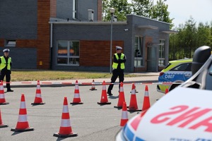 na pierwszym planie fragment ambulansu w tle policjant umundurowany oraz zabudowania