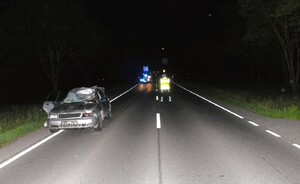 Samochód osobowy marki Audi z uszkodzonym przodem i dachem i szybą czołową stoi n drodze w tle funkcjonariusz policji