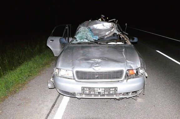Samochód osobowy marki Audi z uszkodzonym przodem i dachem i szybą czołową stoi n drodze w tle funkcjonariusz policji