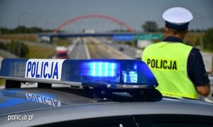 Policjant dach radiowozu z sygnałami a w tle droga szybkiego ruchu z wiaduktem