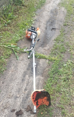 Kosa spalinowa leżąca na drodze gruntowej porośnięta trawą