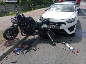 Uszkodzony samochód osobowy koloru białego marki KIA, który jest wbity w uszkodzony motocykl. Pojazdy stoją na asfaltowej jezdni