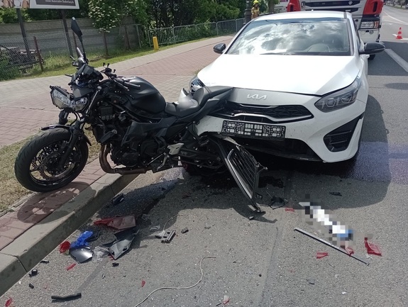 Uszkodzony samochód osobowy koloru białego marki KIA, który jest wbity w uszkodzony motocykl. Pojazdy stoją na asfaltowej jezdni