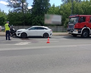 Uszkodzony samochód osobowy koloru białego marki KIA, który jest wbity w uszkodzony motocykl. Pojazdy stoją na asfaltowej jezdni. Obok stoi wóz strażacki i policjant.