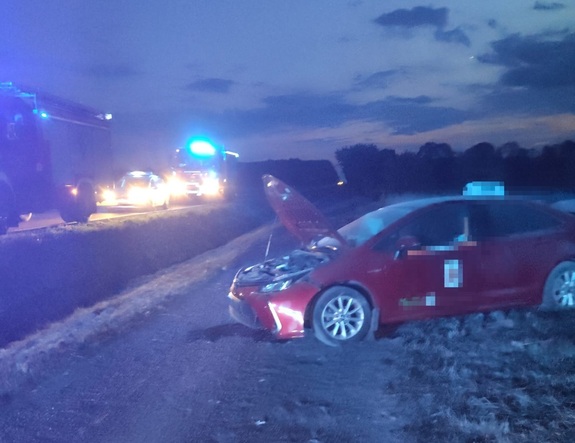 Czerwona osobowa toyota z uszkodzonym przodem stoi na drodze szutrowej i poboczu drogi. Na jezdni widoczne pojazdy słuzb z włączonymi sygnałami.