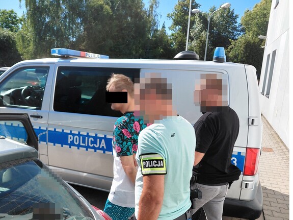 Dwóch policjantów nieumundurowanych prowadzi zatrzymanego mężczyznę z założonymi kajdankami na ręce z tyłu pomiędzy radiowozami policyjnymi.