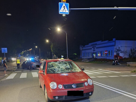 Samochód osobowy koloru czerwonego stojący na ulicy przy przejściu dla pieszych z uszkodzonym przodem w wyniku zdarzania drogowego w tle stoją pojazdy służb - policji