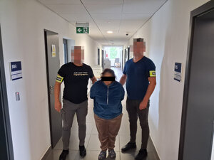 Zatrzymana kobieta  prowadzona korytarzem  przez nieumundurowanych policjantów z opaskami z napisem policja na ramieniu.
