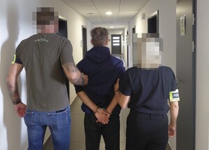Zatrzymany mężczyzna ubrany w bluzę i spodnie ma założone kajdanki na ręce trzymane z tyłu i jest trzymany przez nieumundurowanego policjanta i policjantkę z opaskami z napisem policja.