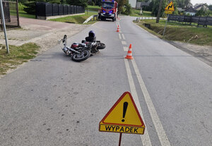 Miejsce wypadku drogowego na jezdni leży motocykl w tle widoczne pojazdy służb - straży pożarnej