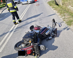 Miejsce wypadku drogowego na jezdni leży motocykl w tle widoczne pojazdy służb - policji i strażak