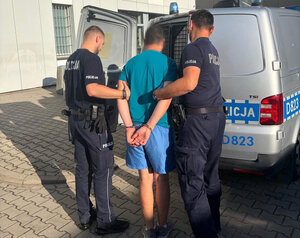 Dwóch policjantów prowadzi zatrzymanego  mężczyznę z kajdankami na rękach do radiowozu typu bus.