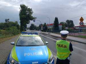 Policjant ruchu drogowego stojący obok radiowozu nadzoruje rejon przejścia dla pieszych przy szkole