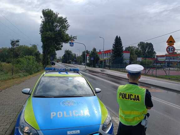 Policjant ruchu drogowego stojący obok radiowozu nadzoruje rejon przejścia dla pieszych przy szkole
