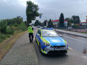 Policjant ruchu drogowego stojący obok radiowozu nadzoruje rejon przejścia dla pieszych przy szkole