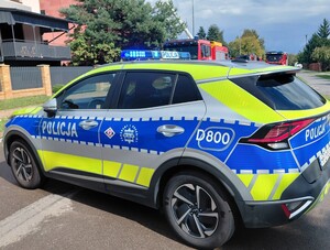 Radiowóz policji z włączonymi sygnałami stojący w poprzek drogi w tle wozy strażackie i zabudowania