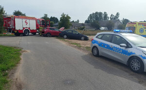 Miejsce zdarzenia drogowego na skrzyżowaniu dróg. Dwa uszkodzone samochody osobowe oraz pojazdy służb dwa wozy strażackie i radiowóz policji. Strażacy stojący obok pojazdów.