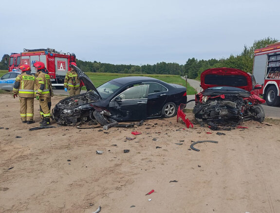 Miejsce zdarzenia drogowego na skrzyżowaniu dróg. Dwa uszkodzone samochody osobowe oraz pojazdy służb dwa wozy strażackie i radiowóz policji. Strażacy stojący obok pojazdów.