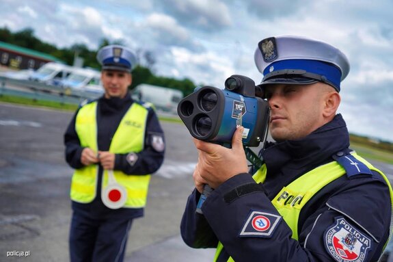 Policjant z mie3nrnikiem prędkości w tle stoi policjantka umundurowana z tarczą