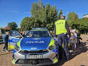 Grupa dzieci stoi na spotkaniu z policjantem w tle radiowóz policyjny