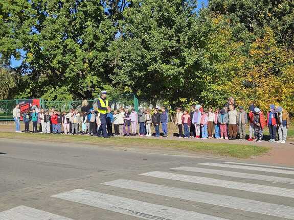 Grupa dzieci stoi na spotkaniu z policjantem w tle radiowóz policyjny