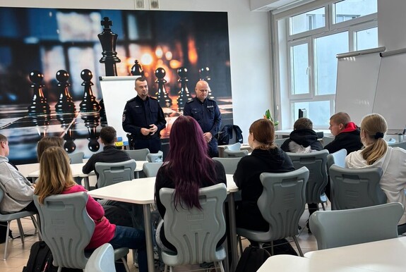 dwóch umundurowanych policjantów prowadzi prelekcję uczniom siedzącym przy stolikach