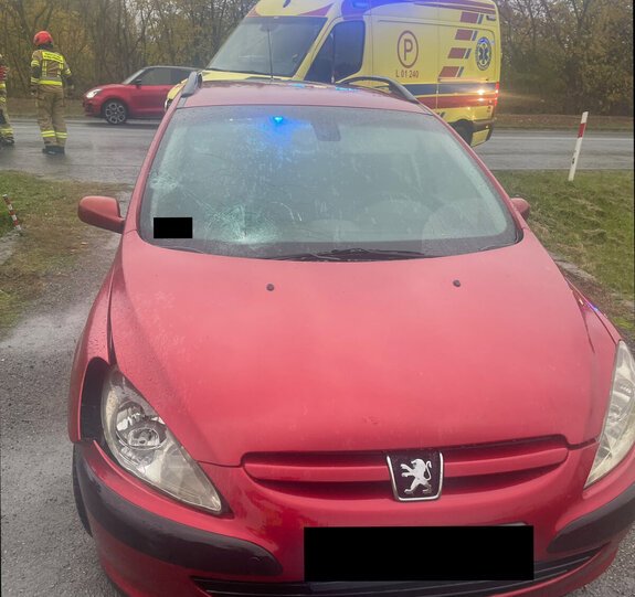 Samochód osobowy Peugeot koloru czerwonego z udszodzoną szyba i błotnikiem w tle karetka pogotowia i droga, strażacy i samochód osobowy