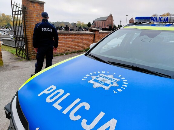 Radiowóz policyjny i policjant stojący przy nim patrzy w stronę cmentarza i kaplicy