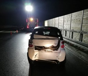 Tył samochodu osobowego marki Smart koloru białego. Uszkodzona tylna cześć pojazdu. Na drugim planie radiowóz straży pożarnej.   Auta stoją na poboczu.